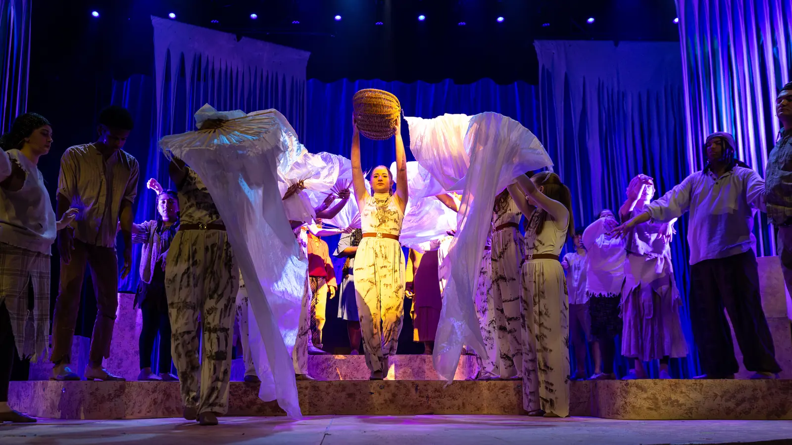 Full company number, the ensemble billowing silks around a soloist lifting a basket overhead